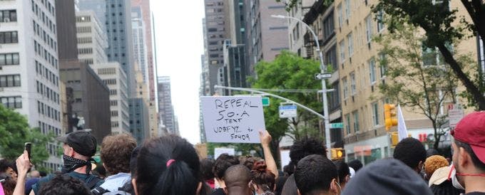 Crowded protest in New York City featuring a protestor holding a sign that reads "Text UOBFYZ to 50409"