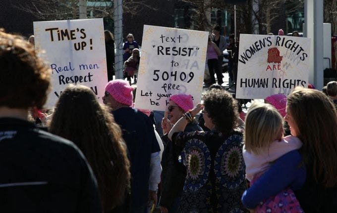 Image of people at a protest, among the protestors are three women in pink hats holding up signs. One reads, "text resist to 50409, free, let your voice be heard."